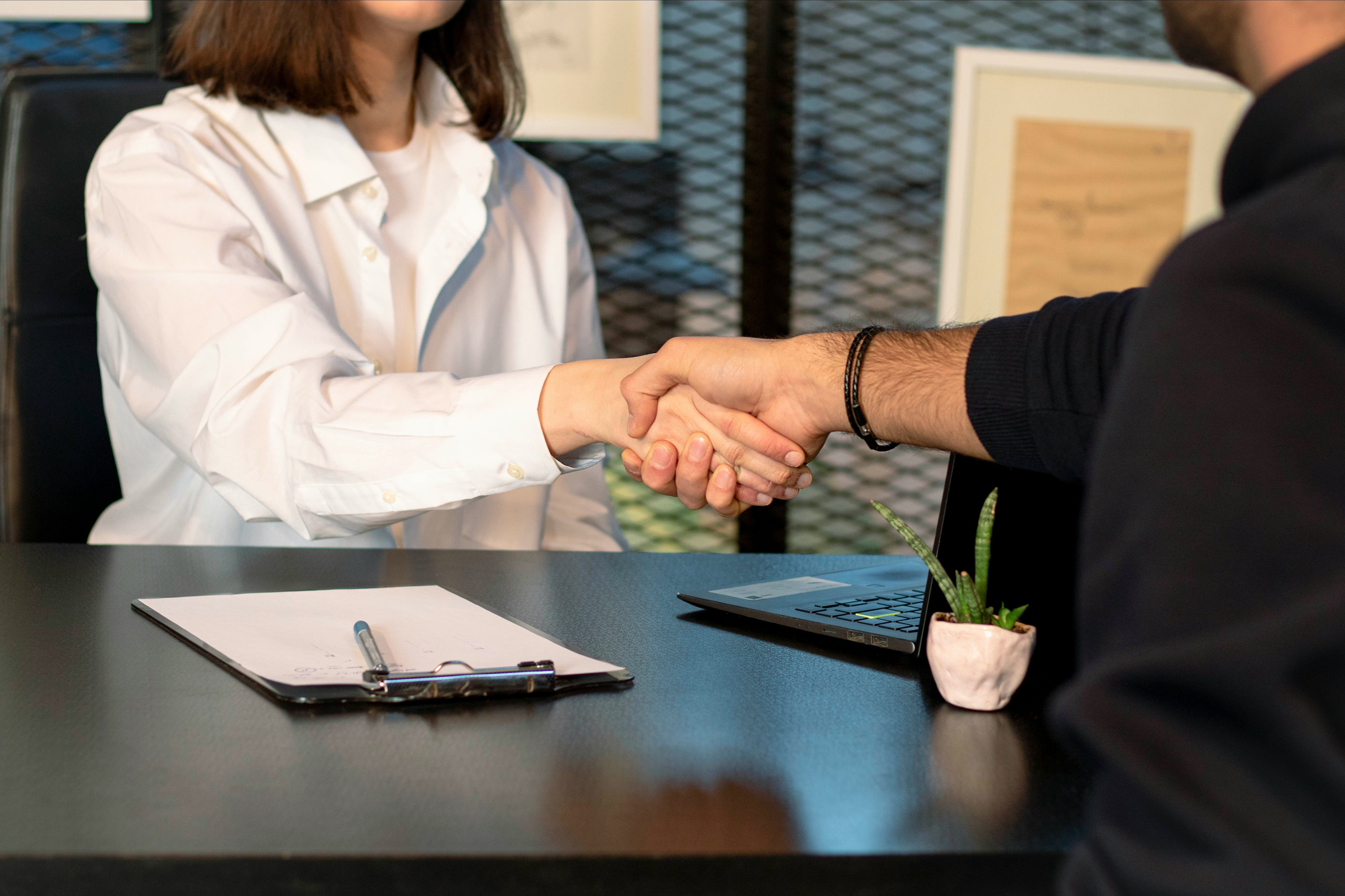 Two people shaking hands across a desk with a clipboard and laptop