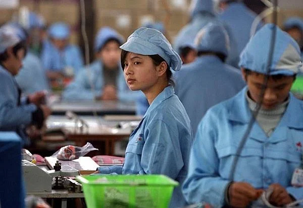 Factory workers in blue uniforms and caps at assembly benches, one looking at the camera