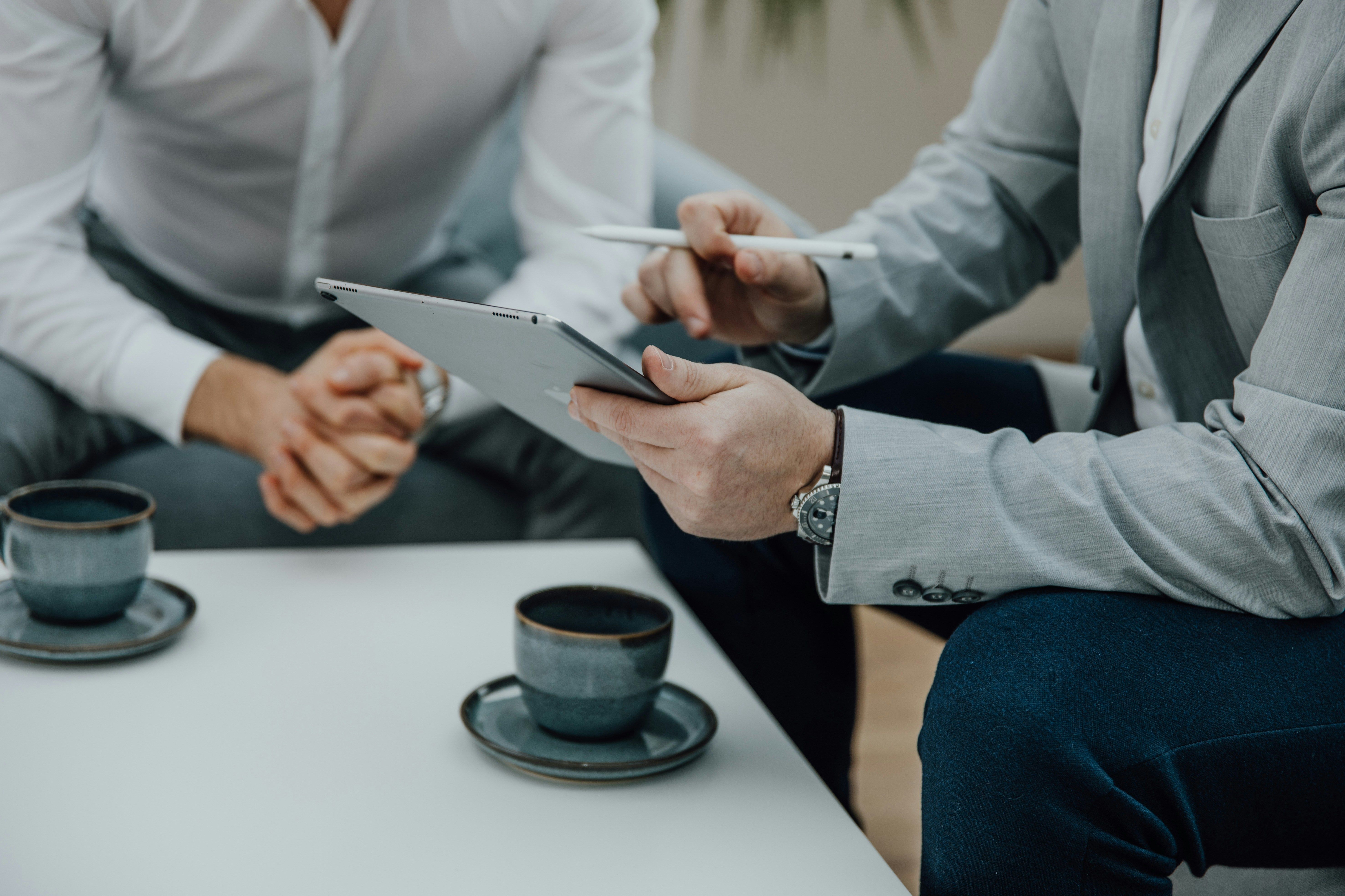 Two businessmen at a low table reviewing a tablet, with two cups of coffee