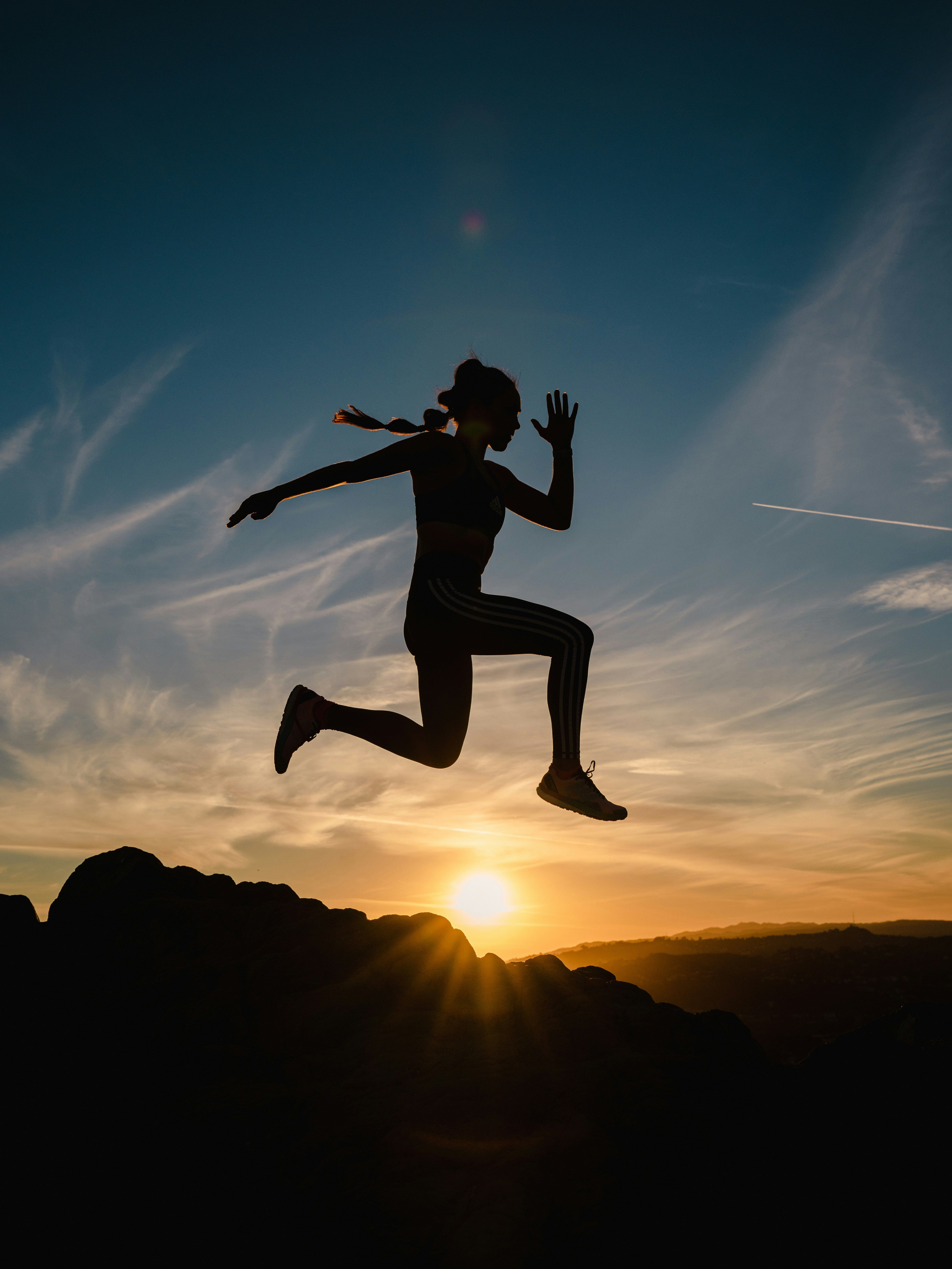 Silhouette of a woman leaping through the air against a sunset sky over dark rocks