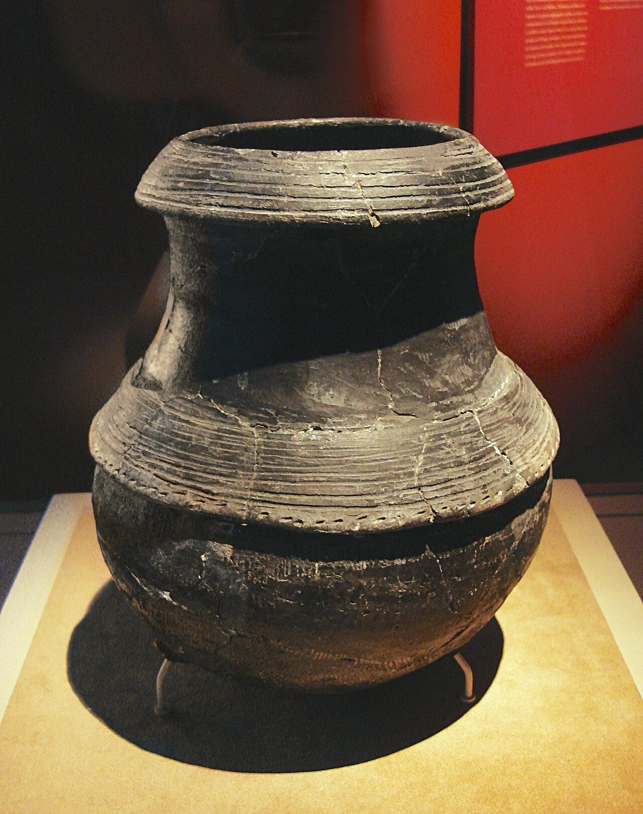 Ancient dark clay pot with ridged bands displayed on a pedestal in a museum