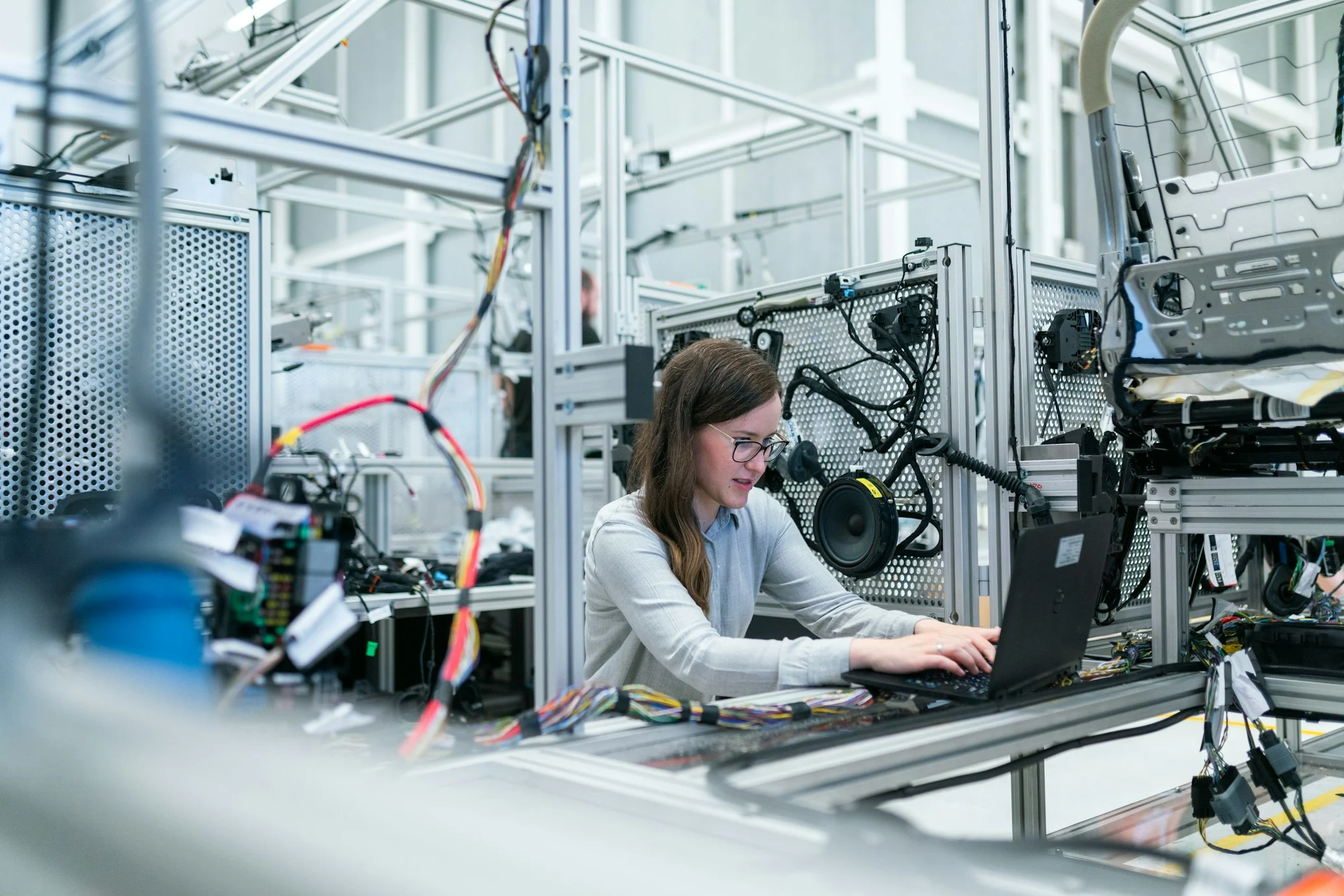 Woman with glasses working on a laptop among wiring and equipment in an electronics workshop